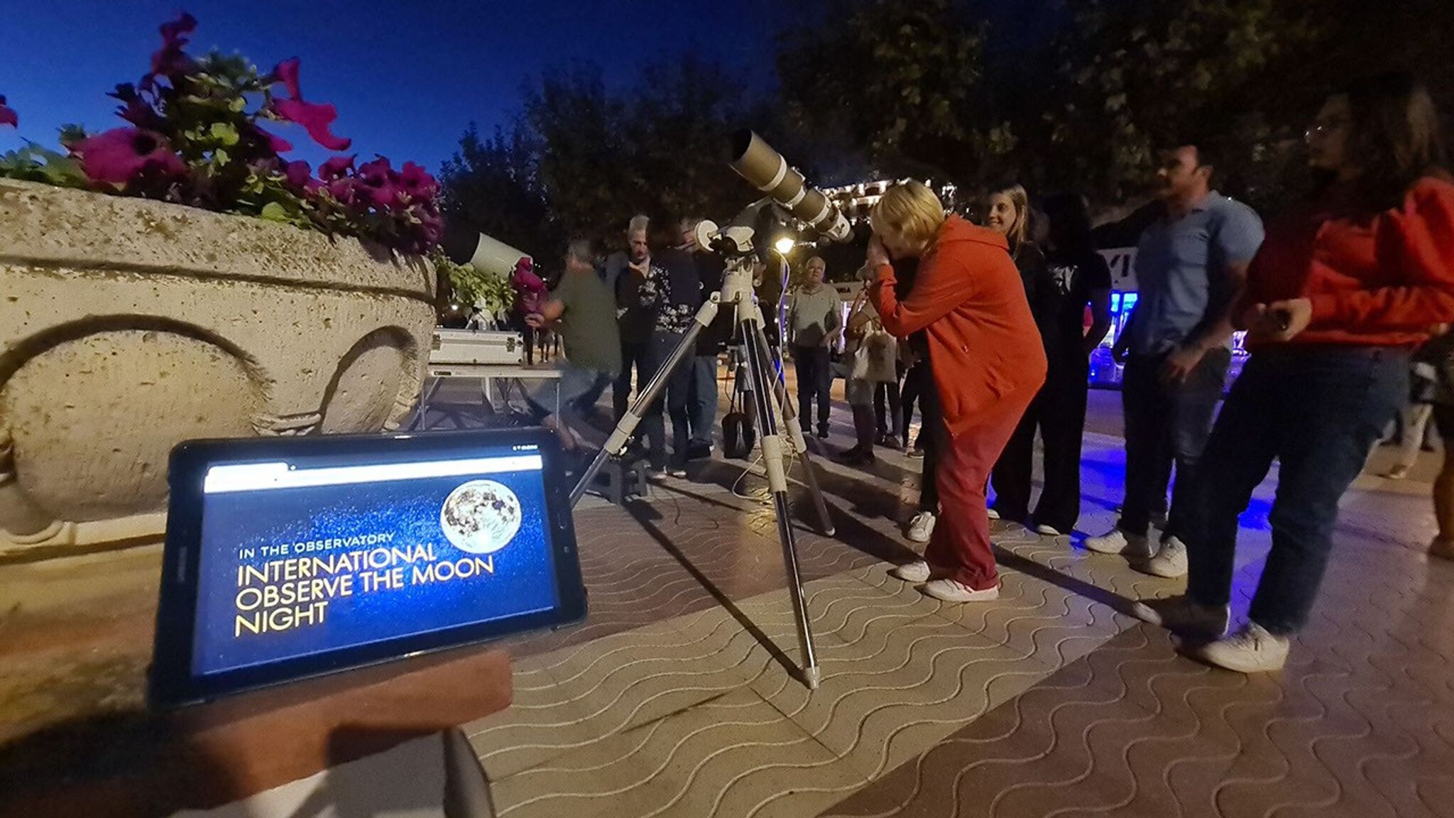 A group of people gathered at night to look through a telescope. In the foreground, a screen displays "International Observe the Moon Night"