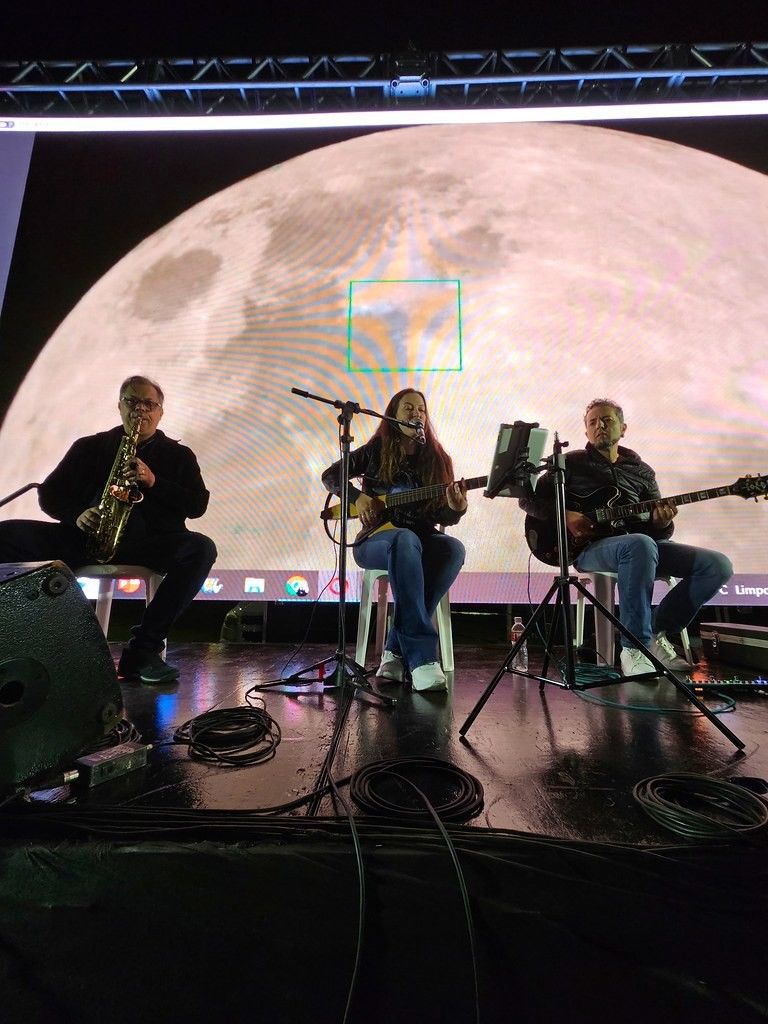Photo of three musicians performing on a stage, with a gigantic Moon shown on a screen behind them.