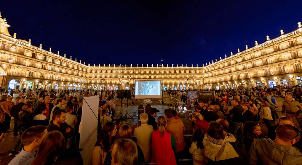 A large crowd gathering at night in a town square watches a screen with an image of the Moon surface.