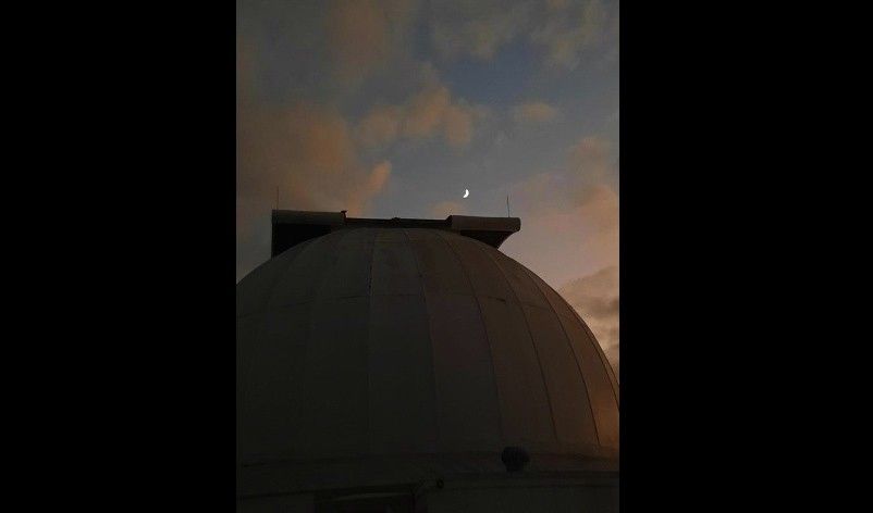 Photo of the evening sky with a tiny crescent Moon in the sky and clouds above a domed building structure.