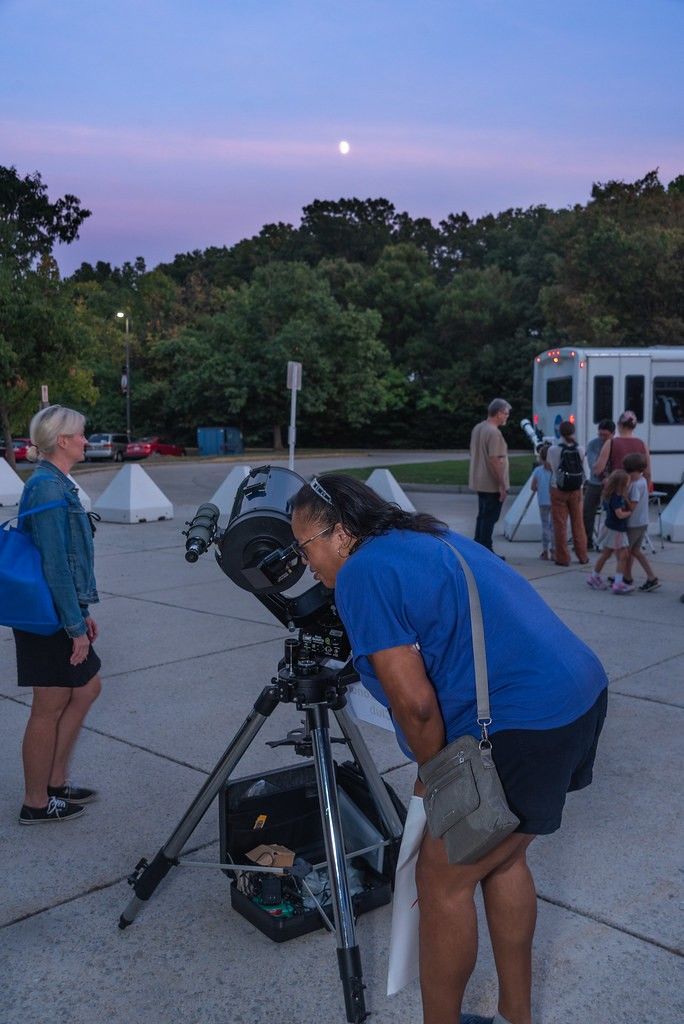 A person wearing a blue T-shirt peers through a telescope pointed at the Moon in the sky.