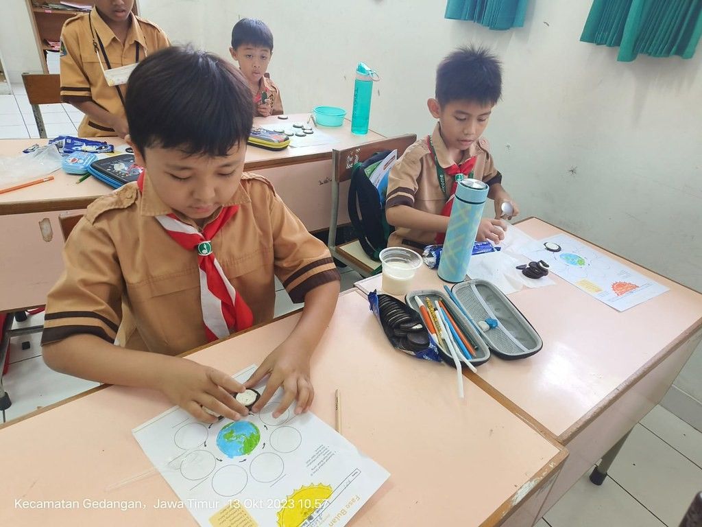 Two young students in tan uniforms with red scarves sit at tan desks with printed sheets on top and colored pencils. The sheets show the Earth in the center with blank circles around it representing the Moon. The student on the left is placing a creme cookie on one of the circles.