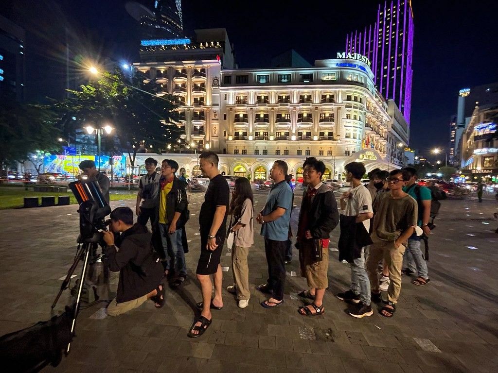 A group of people gathered at night, waiting in line in a city plaza to look through a telescope.