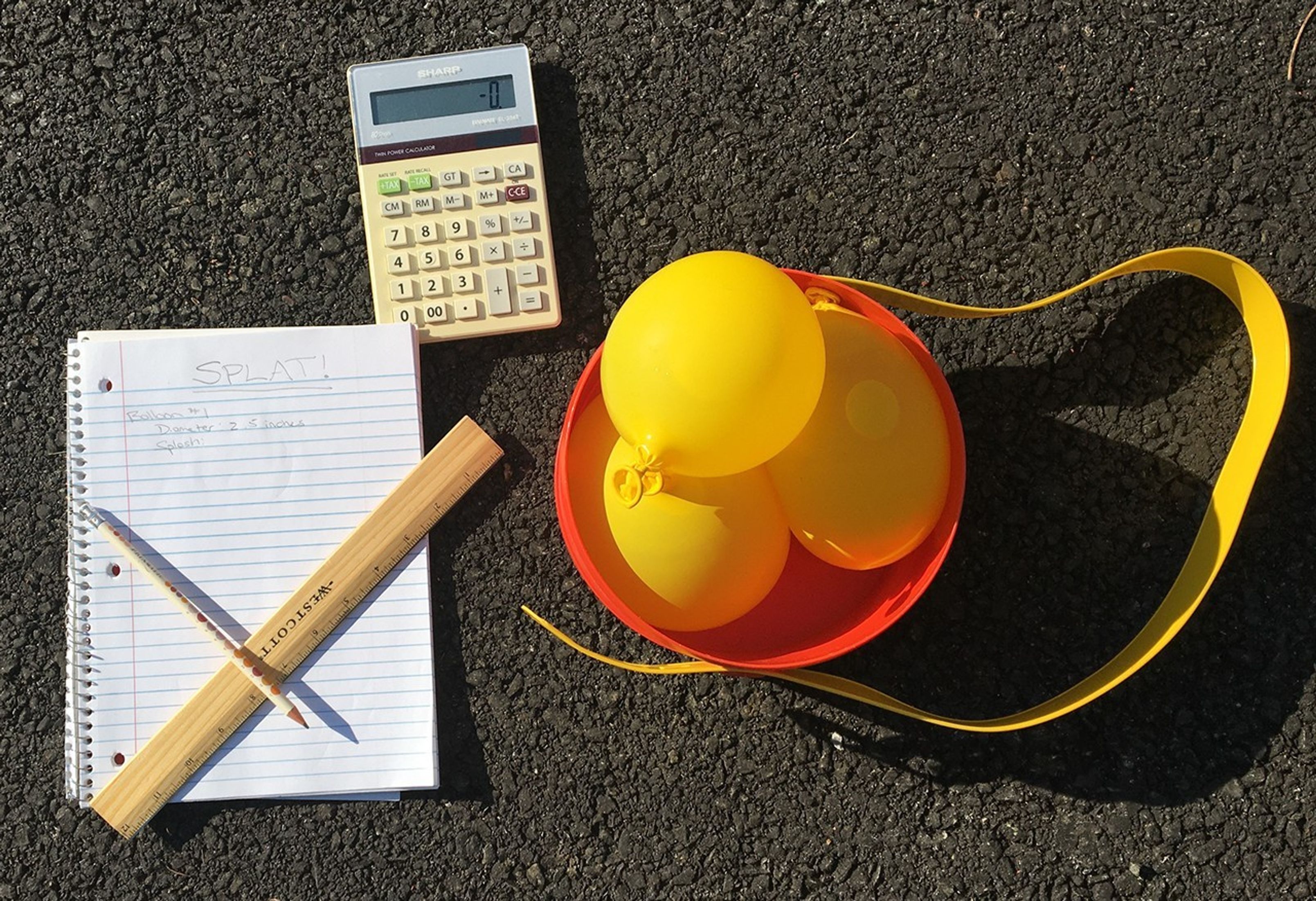 A ruler and pencil are positioned on top of a lined notebook. A calculator and three yellow water balloons are next to the notebook. All materials are lying on top of blacktop.