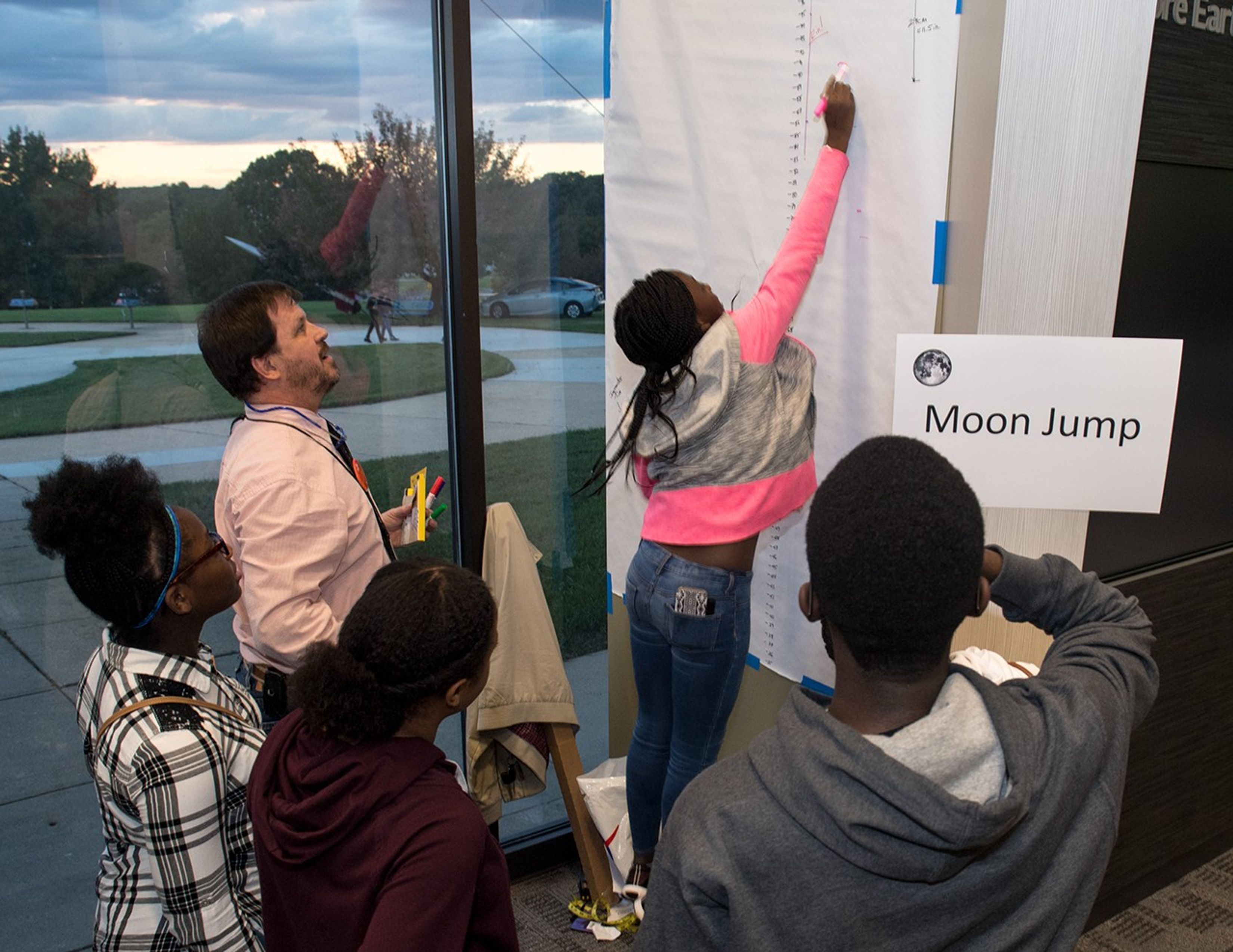 A young woman jumps with a pink marker in her hand, making a mark on a long white roll of chart paper. Four others watch on around her.