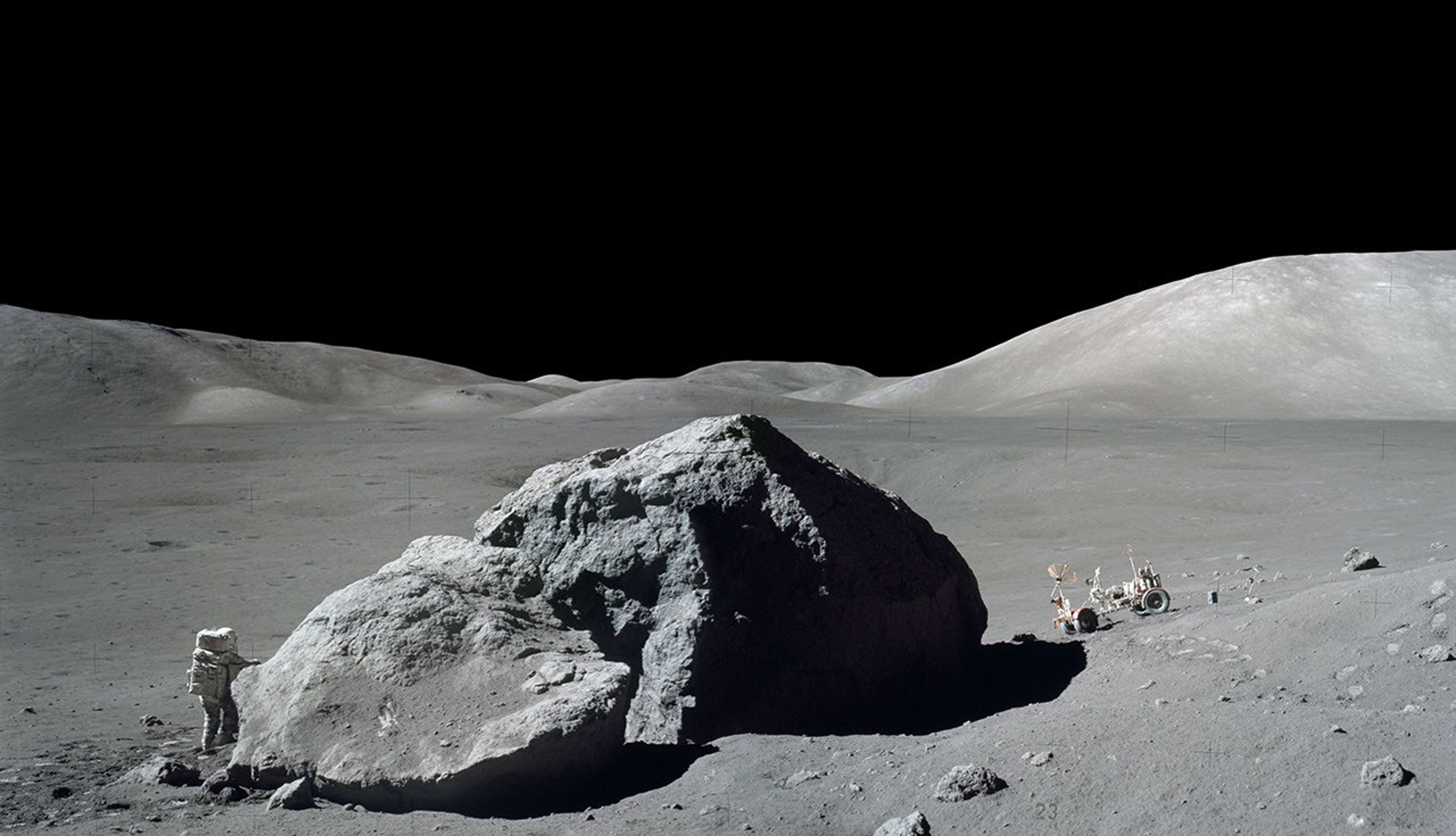 Astronaut and rover on the Moon's surface, separated by a boulder several times taller than the man.