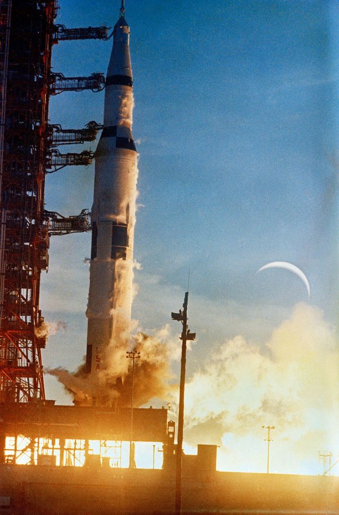 Saturn V rocket launches with great flames, with the Moon in the sky in the background