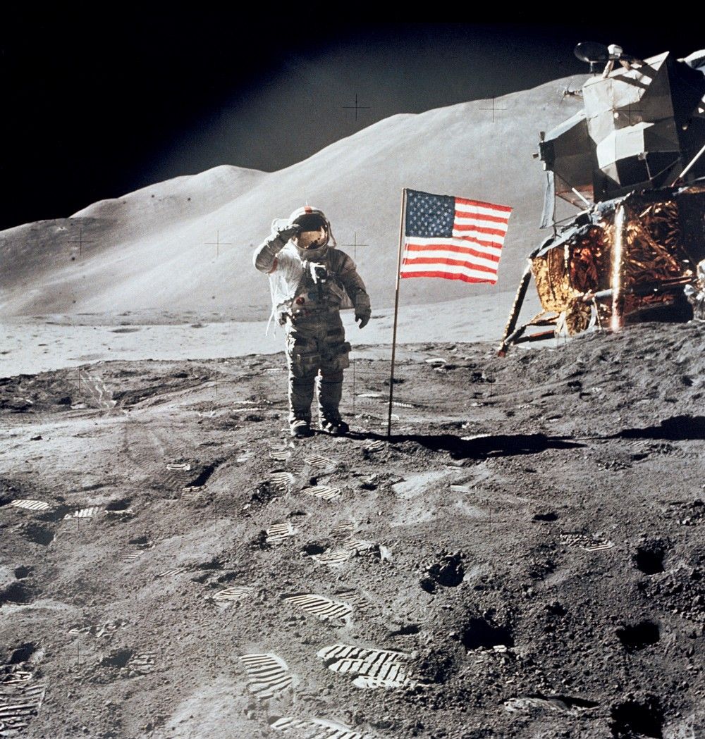 Astronaut giving salute beside American flag on the Moon, with lander in the background. In the foreground: bootprints in fine, grey dust.