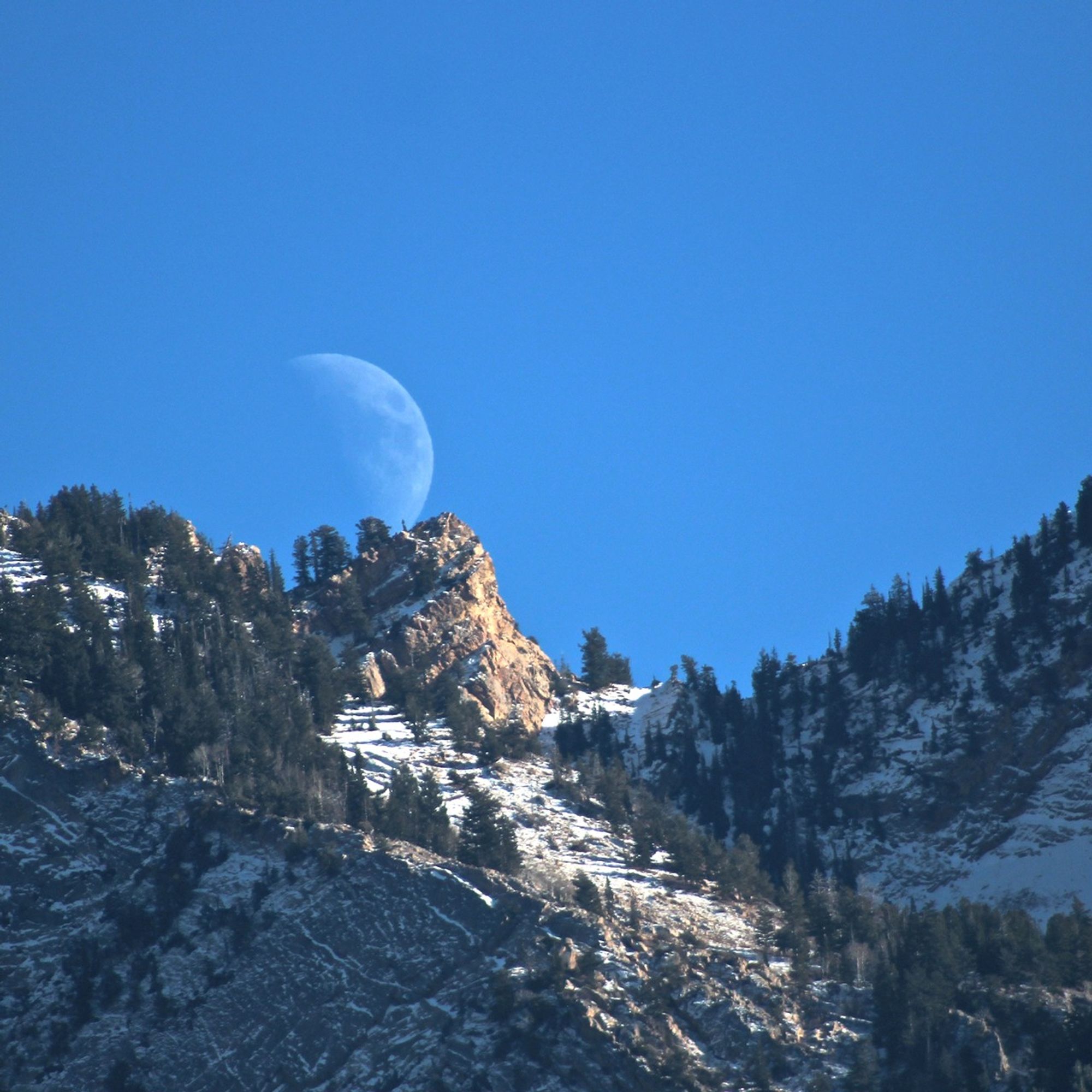 A faint waning Moon rises in daylight over rugged, snowy mountains