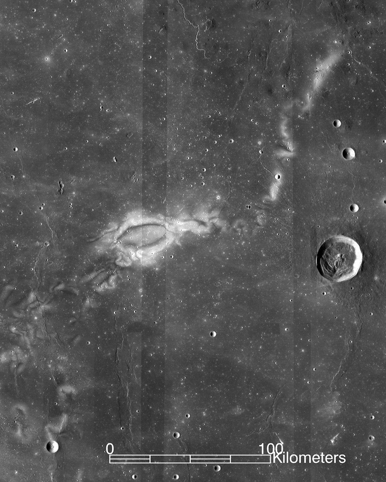 A white curly eye-like shape over a gray-scale pock-marked surface. Satellite image of the Moon. A scale bar shows the image is over 100 kilometers wide.