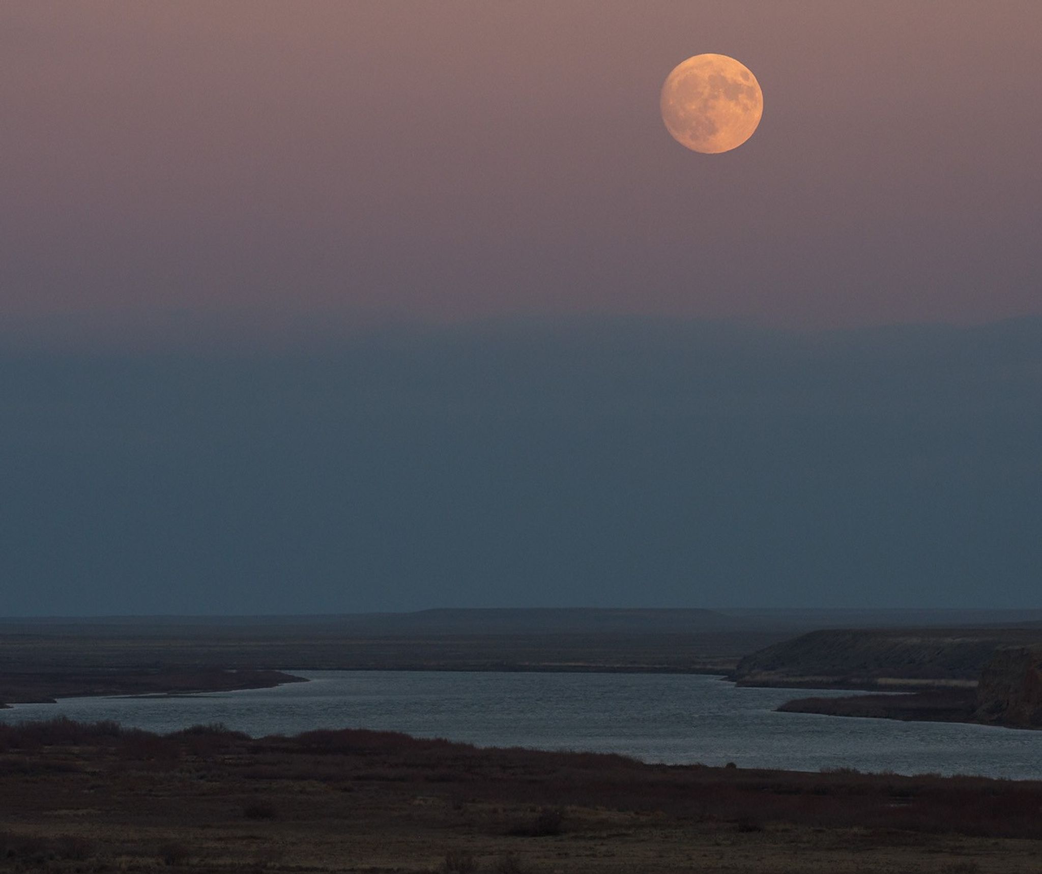 At twilight, a full moon rises over a broad river with vegetated banks. The Moon appears peach-colored in a dim pink-and-blue sky.