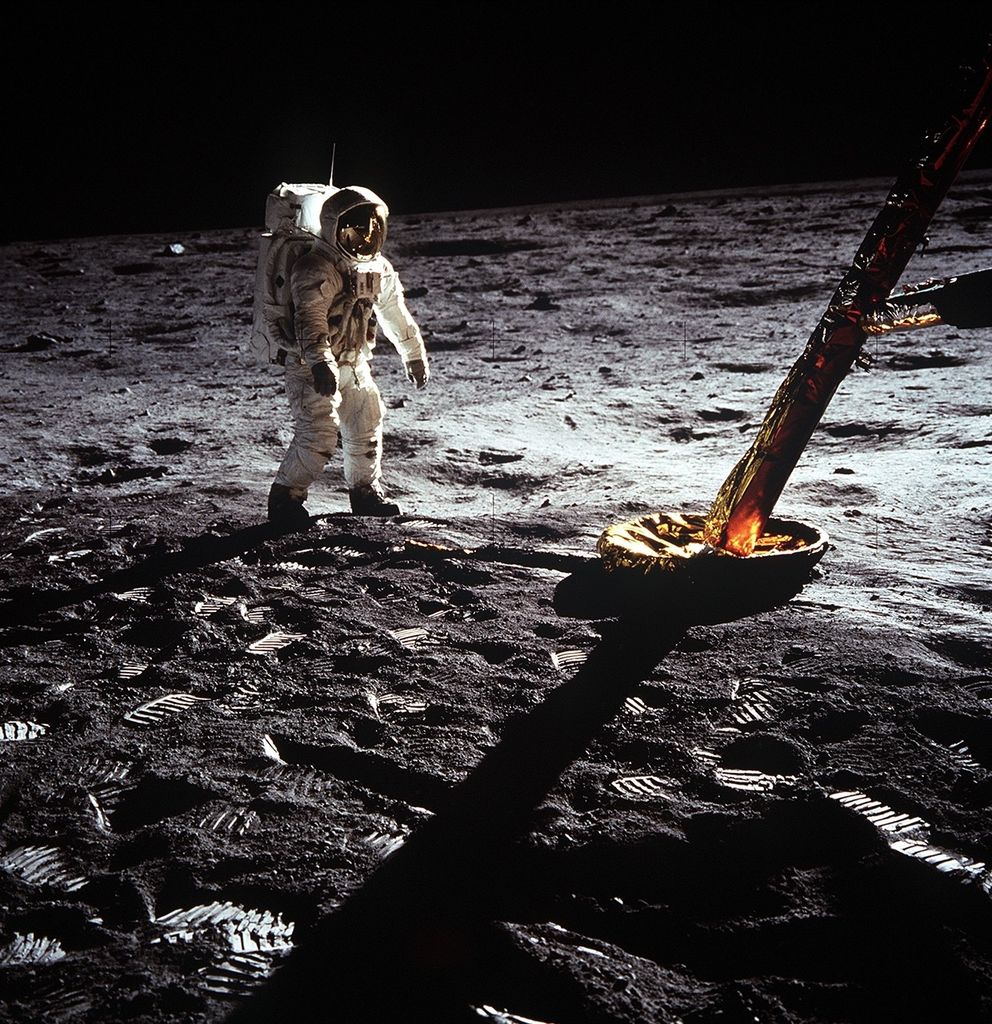 Astronaut on the Moon near the landing gear of the lunar module, standing on a surface covered with footprints and lunar dust, under a pitch-black sky.