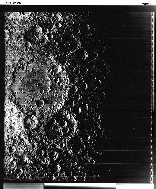 A black and white image of overlapping craters. Two significantly large craters overlap each other to the left of the image and are pocked with numerous other, smaller craters. The right side of the image is in shadow and black.