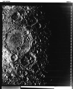 A black and white image of overlapping craters. Two significantly large craters overlap each other to the left of the image and are pocked with numerous other, smaller craters. The right side of the image is in shadow and black.