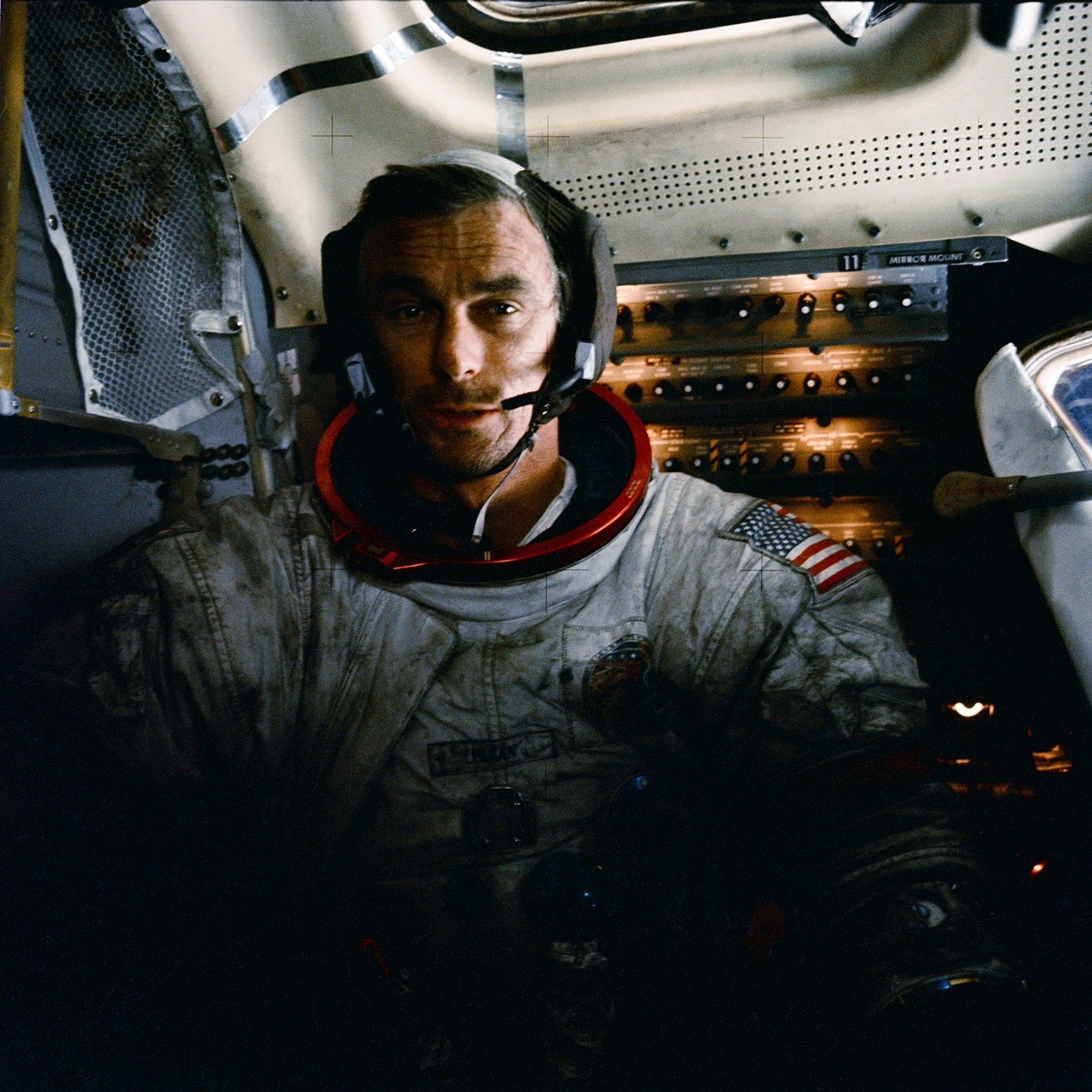 An astronaut sitting inside the Apollo command module spacecraft with his helmet off. He wears a microphone and his white spacesuit appears dusty.  