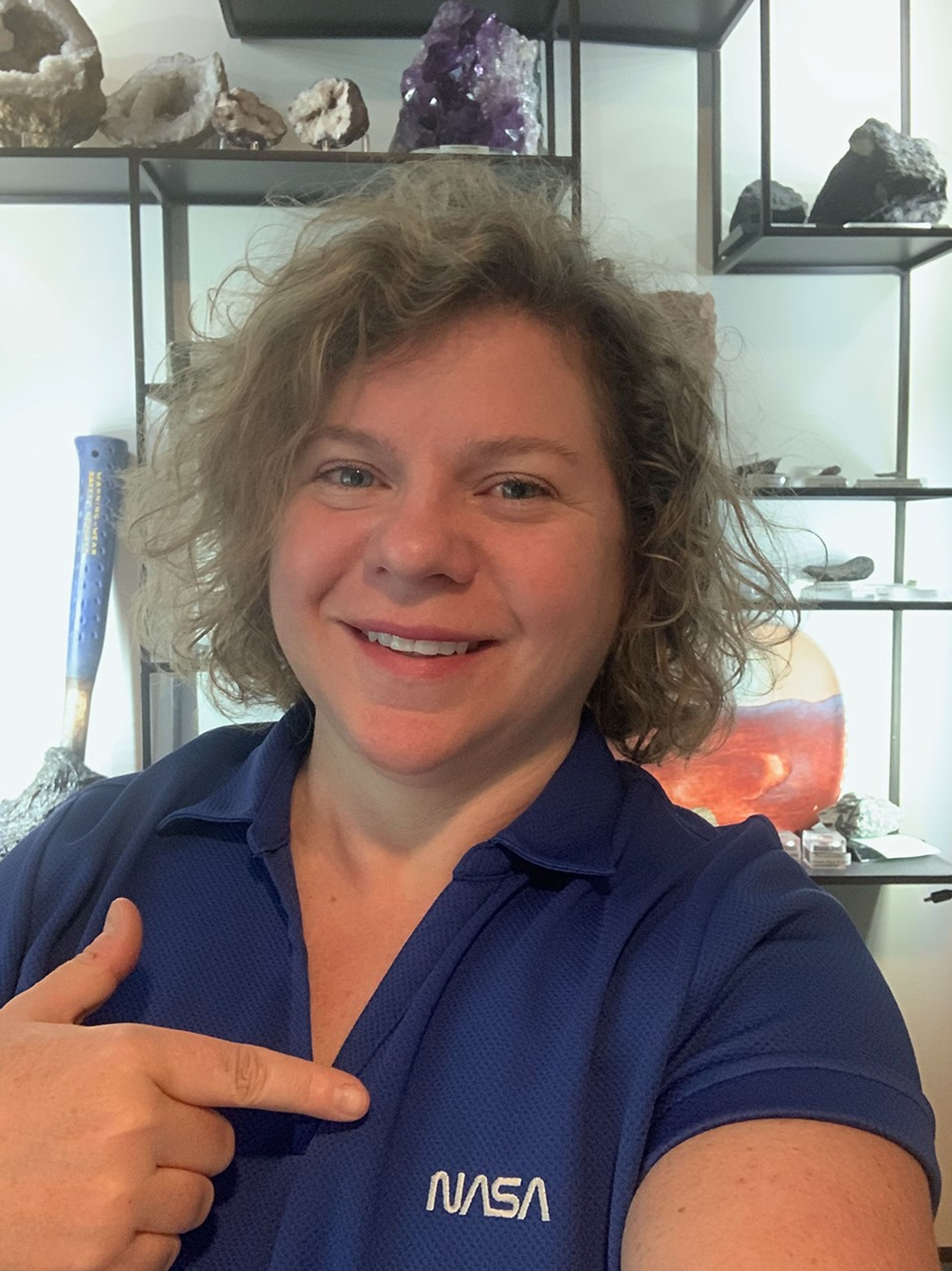 A portrait photo of NASA scientist Barbara Cohen. Barbara wears a blue NASA polo shirt and points to the logo. Barbara stands in front of shelves with various crystals and rocks.