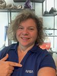 A portrait photo of NASA scientist Barbara Cohen. Barbara wears a blue NASA polo shirt and points to the logo. Barbara stands in front of shelves with various crystals and rocks.