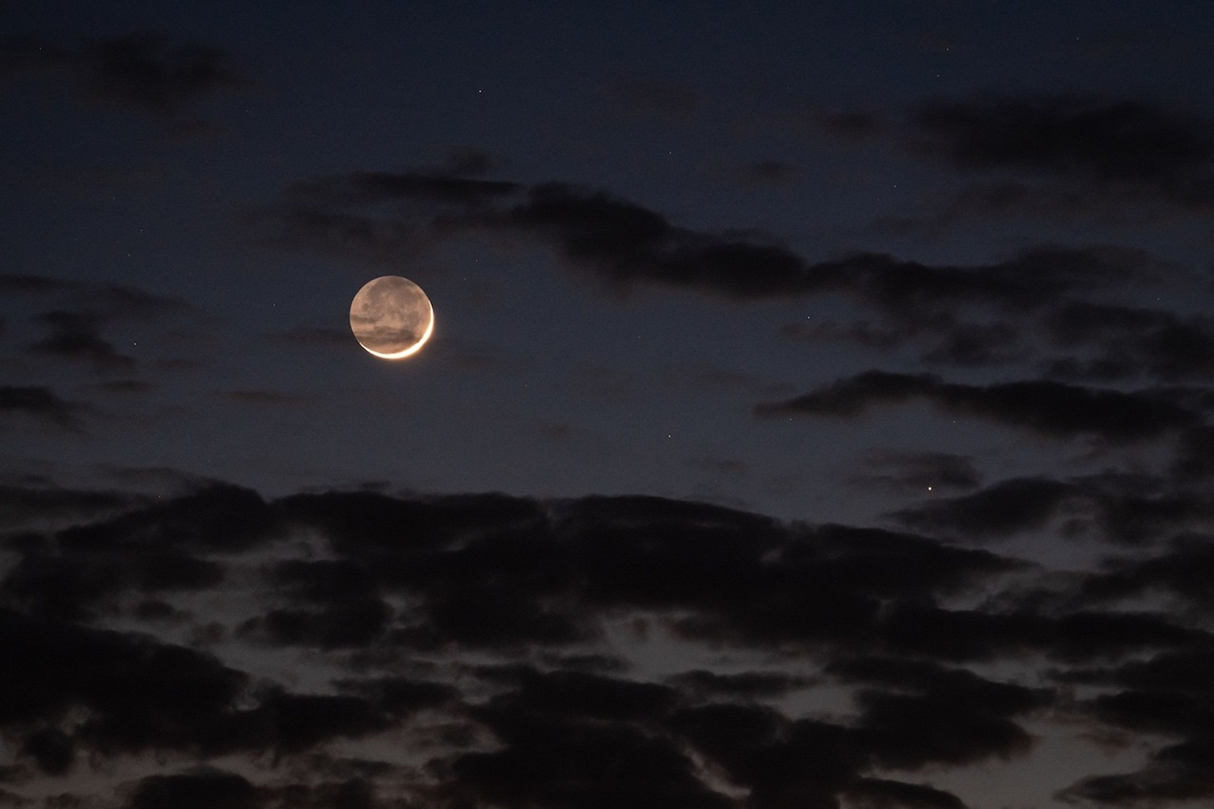 Photo of Moon in a dark, cloudy sky. Though the Moon is in a crescent phase, its whole near side is also dimly visible.