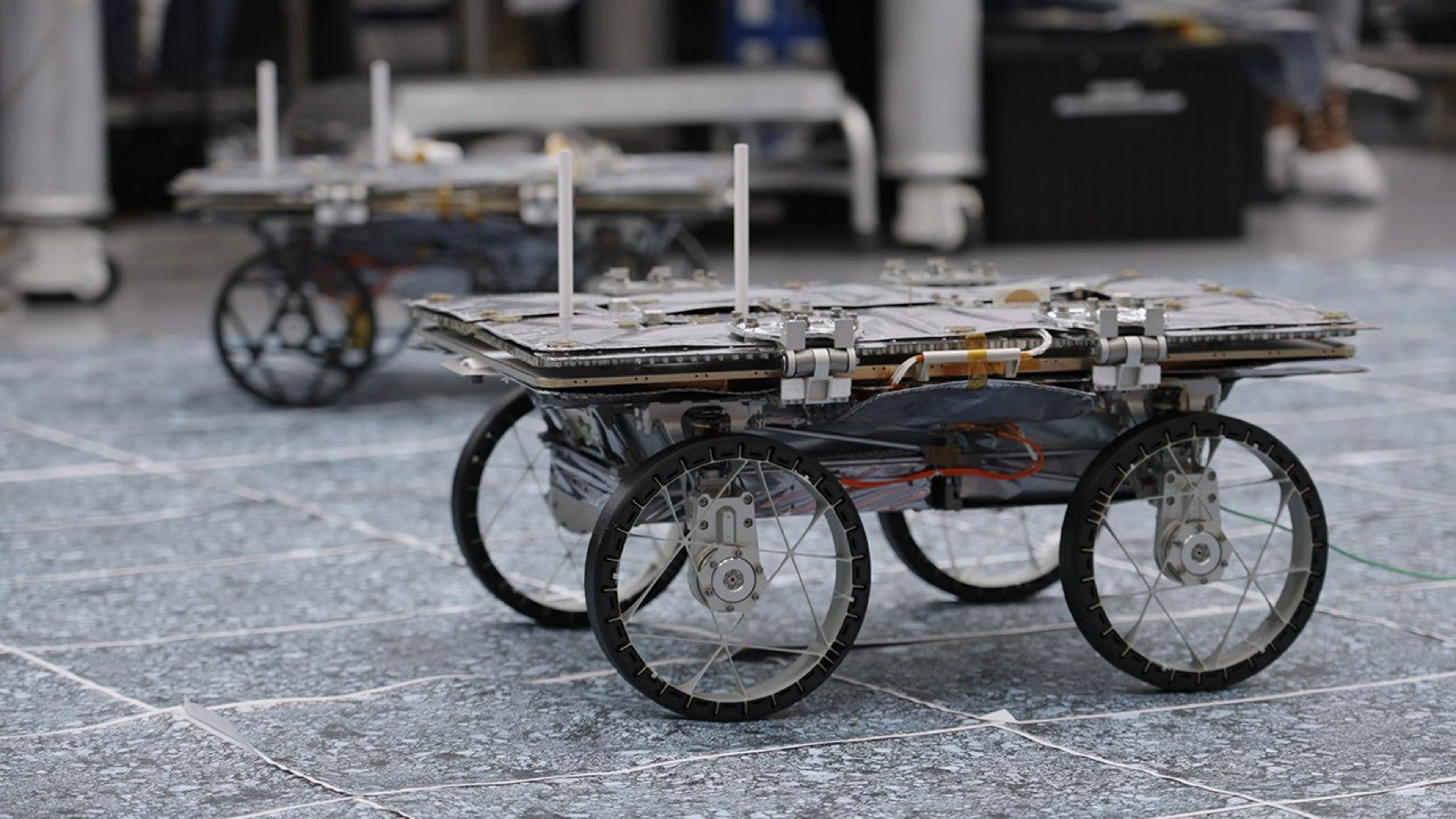 A suitcase-sized robot on 4 wheels sits on the floor in a lab. Wires run across the body. The top of the robot is flat except for 2 antennae. There is a similar robot in the background.