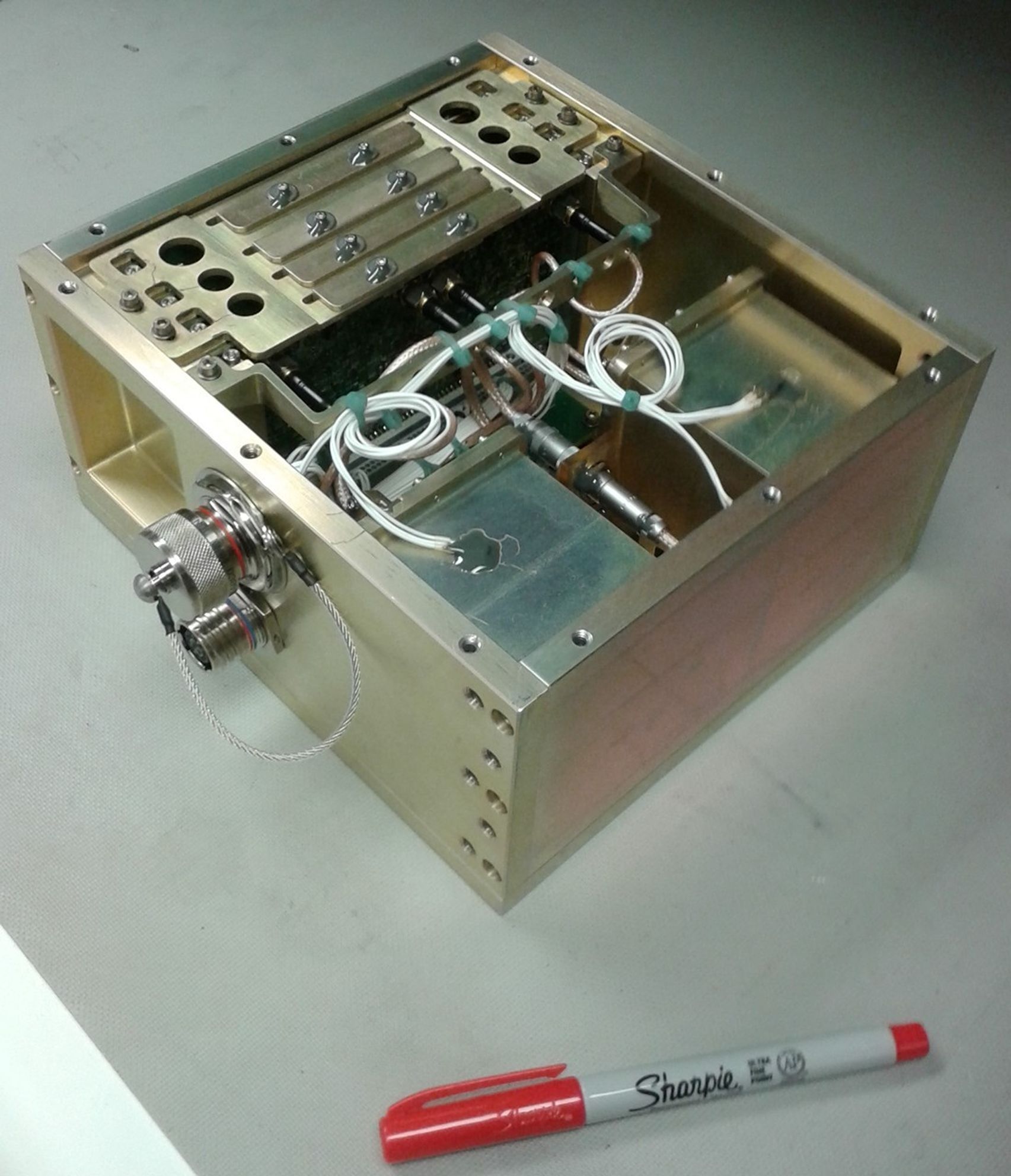 An image of a sealed Neutron Measurement System on the surface of a table. A sharpie sits next to it for scale. The outer layer is reflective. The size is about 5 inches by 7 inches by 7 inches.