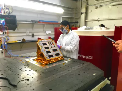 A scientist wearing protective equipment works on an array of solar panels in a laboratory. There are 8-10 different solar panels next to one another.