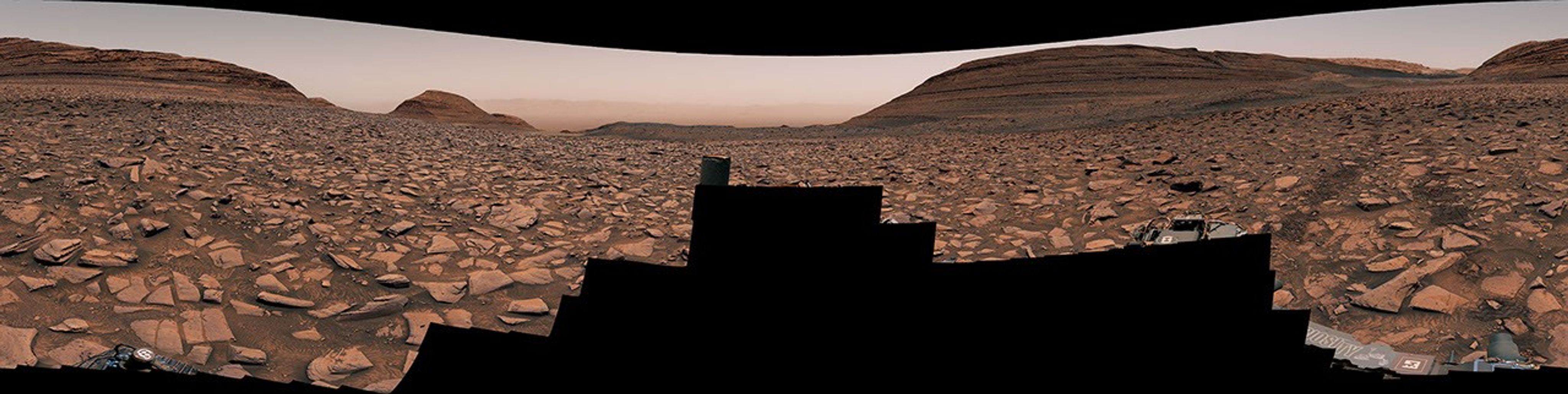 A rocky plain extends to the horizon dominated by small foothiils. The rover tracks are visible in the soil.