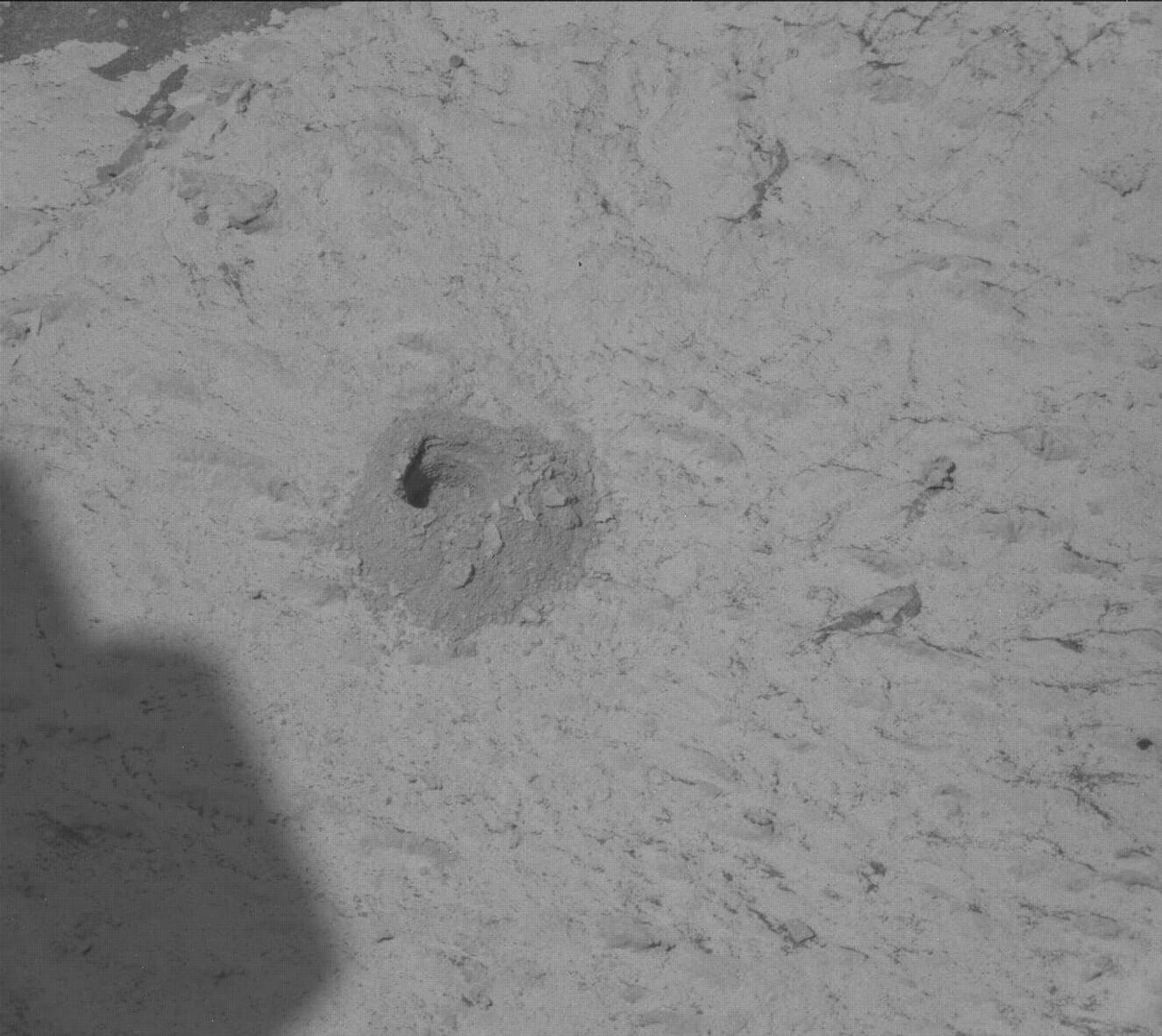 This black and white image shows a quarter-size drill hole on the rocky, sandy surface of Mars. Curiosity's shadow is also present in the bottom left corner of the image.