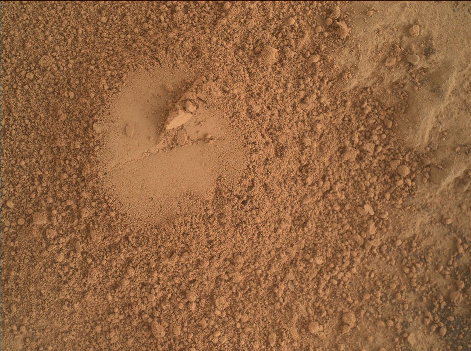 This is a colored image of sand and rock scattered on a smooth surface. On the center of the image, a smooth circular surface is present.