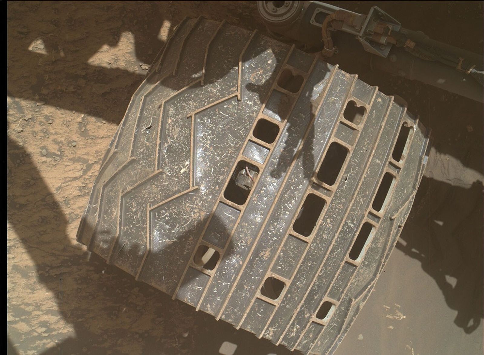 This is a large colored and zoomed in image of one of the wheels on Curiosity. The wheel is located on the sandy surface of Mars. Curiosity's shadow is reflected on the surface.