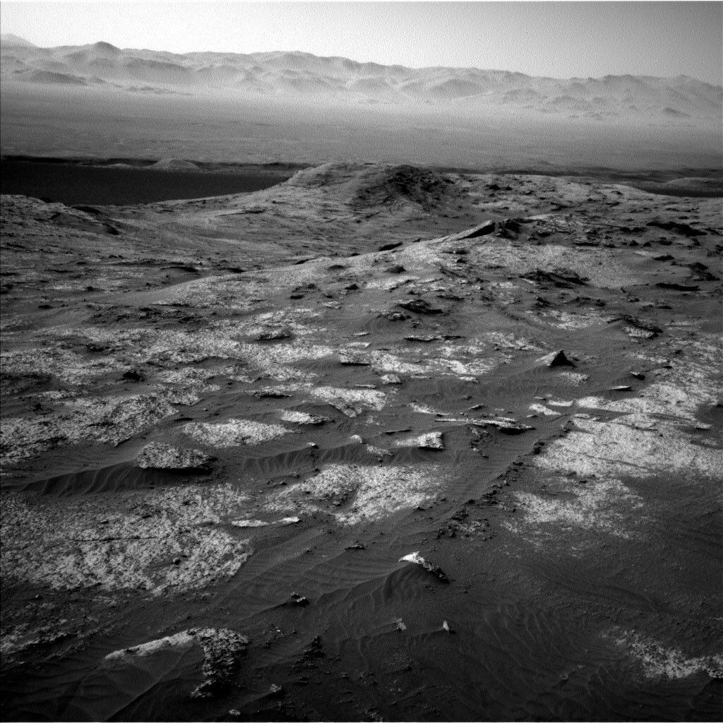 This is a black and white image of the rocky, sandy surface on Mars. In the front of the image rocks are seen embedded into the ground and are slightly poking out in places. Larger hills can be seen in the horizon with a clear sky.