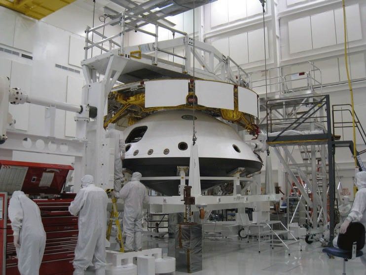 This image shows the cruise structure (the 'ring' on top) and one part of the aeroshell (the backshell with the rover inside) flipped over and the bottom of the aeroshell (the black heat shield) being added. There are four mission team members in the foreground.