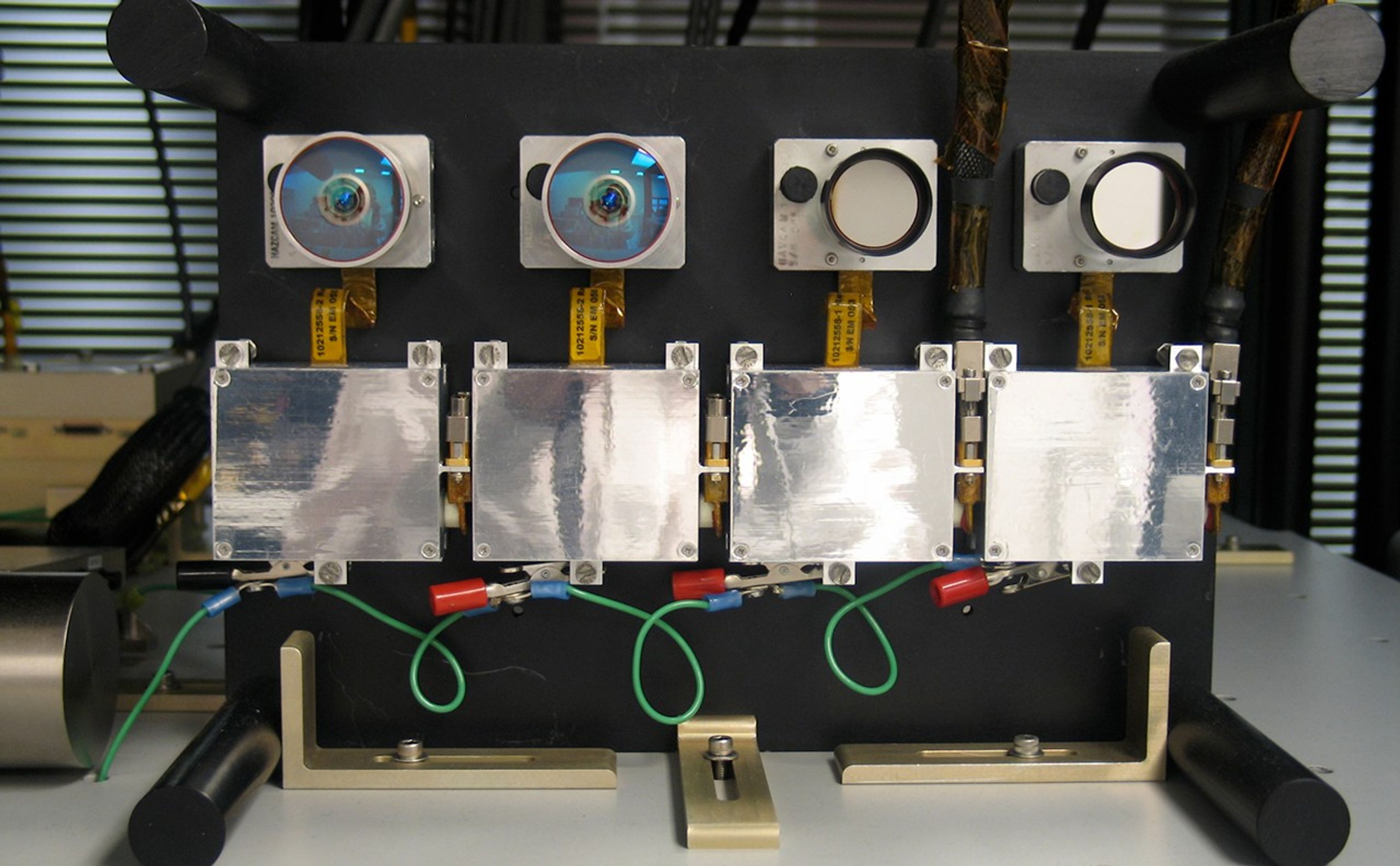 This image shows a closeup view of the two pairs of camera lenses on a shelf. The left-hand pair are larger than the right-hand pair, go to the hazard avoidance camera, and have a larger, fish-eye lens field of view for examining the rover's wheels and immediate surroundings. Its lenses are a clear, almost-turquoise blue. The right-hand pair of lenses are smaller and go to the navigation camera, which takes panoramic, far-field images of surrounding terrain.