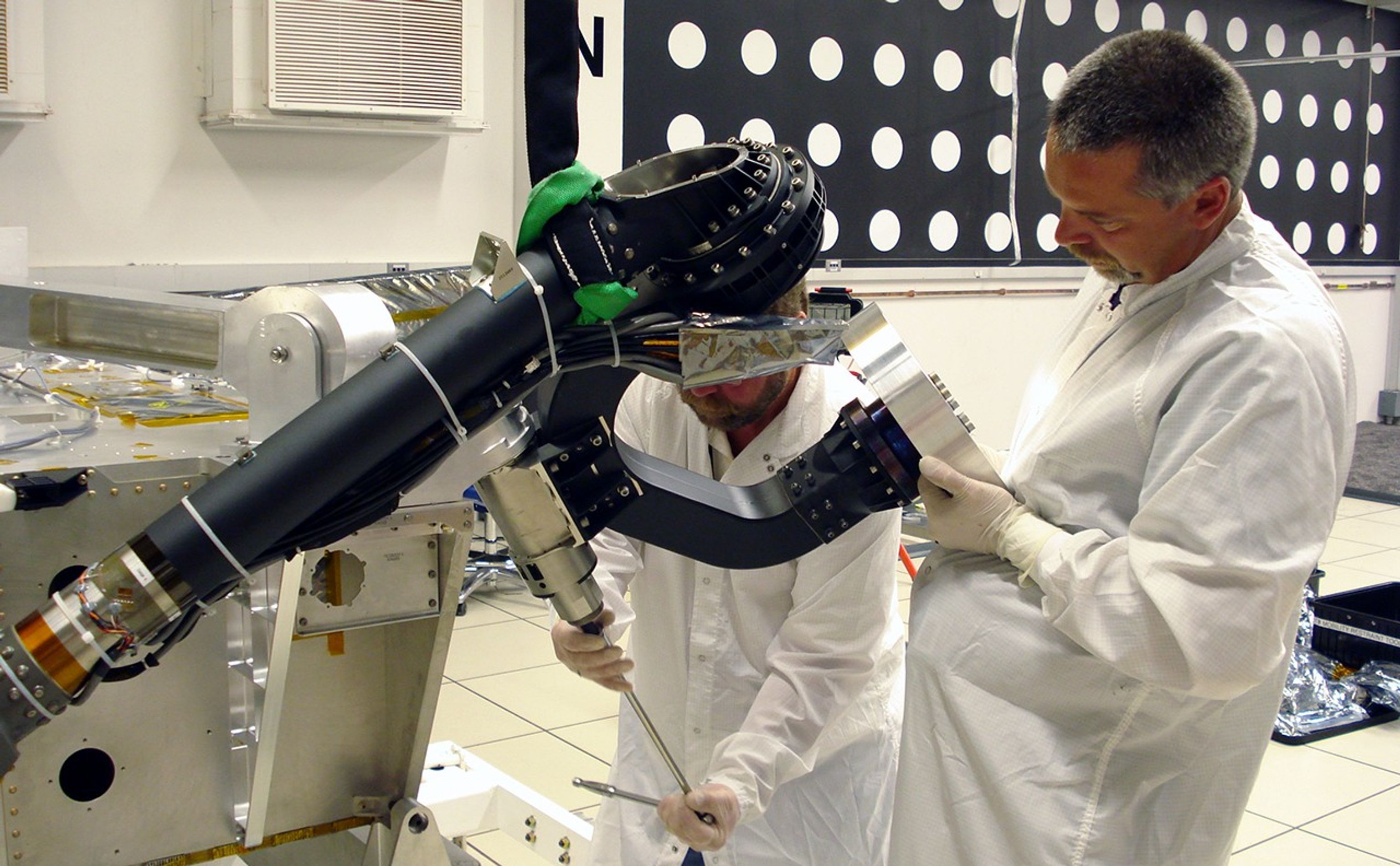 In this image, two technicians from the Mars Science Laboratory team prepare the rover's mobility system for instrumentation tests. They are both wearing white coats and gloves as they each work on one of the rover's 'legs' in a white-tiled room.