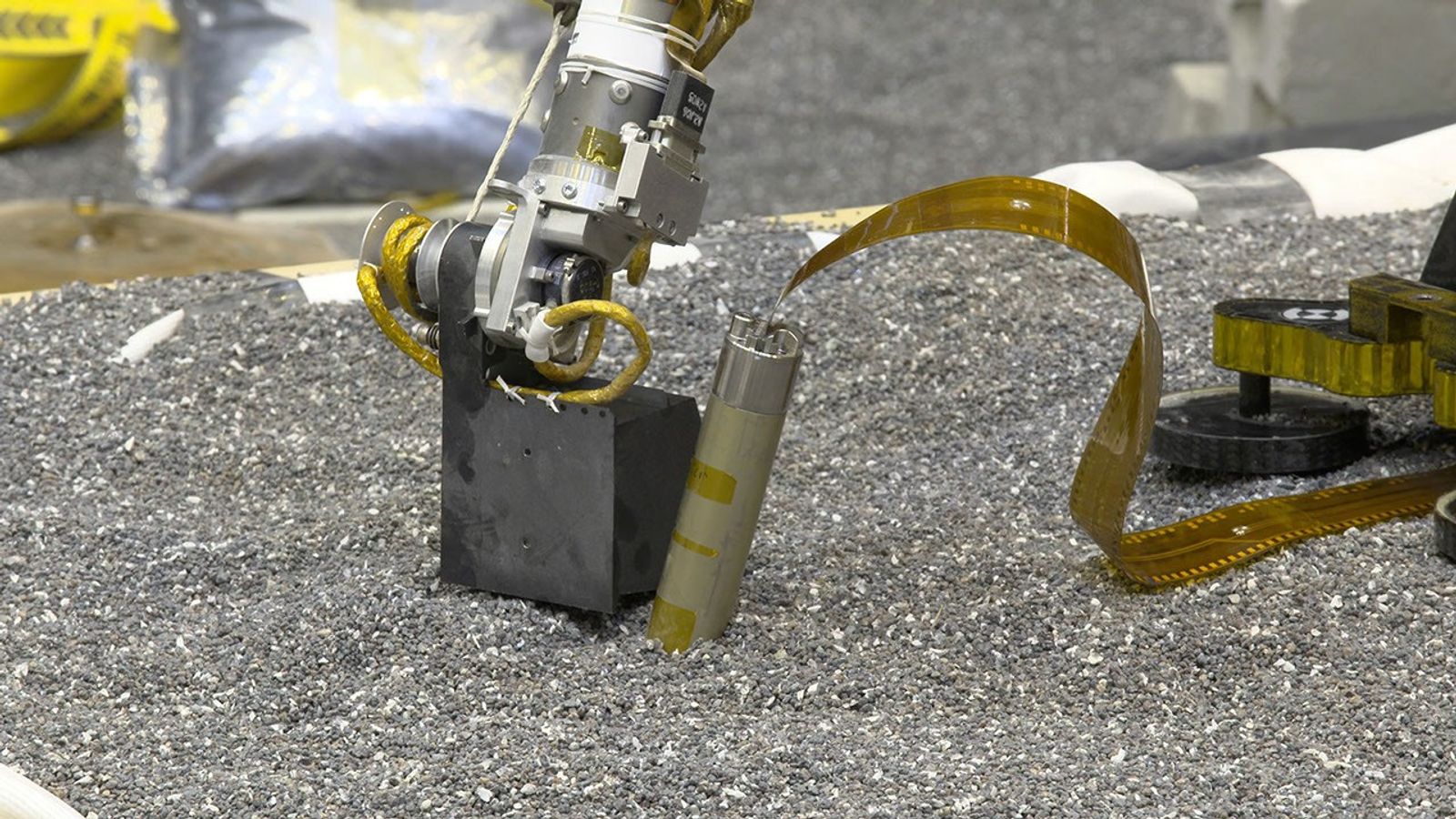 In a JPL lab, a replica of NASA InSight's robotic arm presses with its scoop on crushed garnet near a replica of the spacecraft's self-hammering "mole." Engineers believe pressing like this on Martian soil may help the mole dig by increasing friction of the surrounding soil.