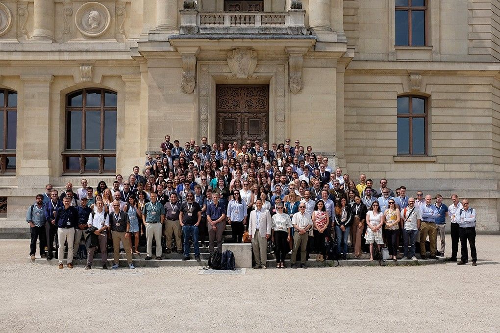 A large group of people, the Mars 2020 Perseverance Rover Science Team, are gathered for a group photo on the steps and in front of a grand, light-colored stone building. The building features large, dark-paned windows and an ornate wooden door at the top of the steps. The people are diverse in age and gender, dressed in a variety of casual to semi-formal attire, with many wearing sunglasses. They are arranged in rows, filling the width of the steps and extending onto the ground in front of them. The sky is bright, suggesting a sunny day.