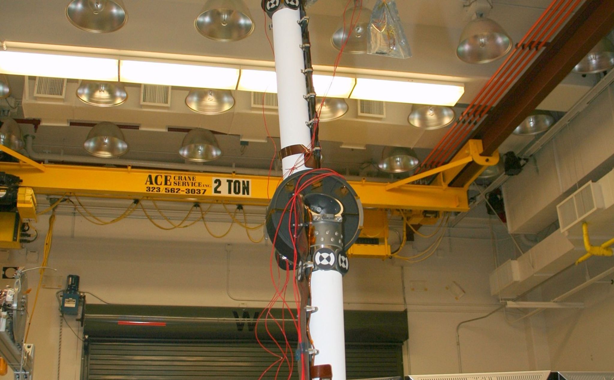 In the middle of this image, the long robotic arm rises straight up toward the ceiling of the lab where it is being tested. The arm is white, and is flexible at several gray round 'joints.' Red wires dangle and are strung along various parts of the arm. At the end of the arm is a complicated set of instruments. In the background, a yellow crane for lifting equipment up to 2 tons (4,000 pounds) can slide along the ceiling to place heavy equipment anywhere in the room. A metal garage-like door and computer stations lie behind the area where the arm is extended.