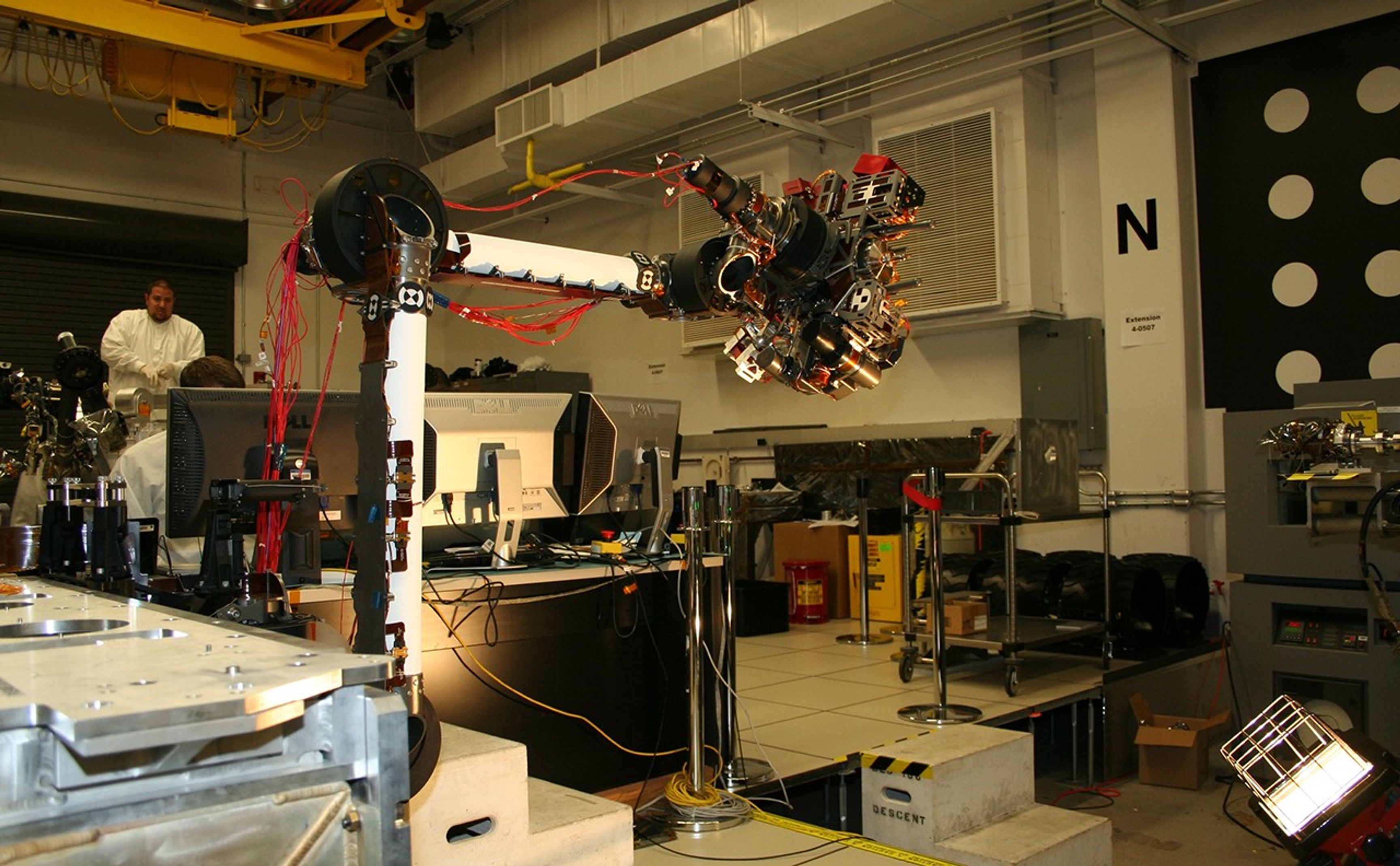 In the middle of this picture, the robotic arm is bent at nearly a 90-degree angle, with the instruments on the end of the arm reaching to the right. Behind the arm is the laboratory where it is being tested. One engineer is hidden behind a bank of computers, while another with a goatee stands watching in the back, beneath a yellow ceiling crane. Both engineers wear white lab coats. Metal lab equipment is scattered throughout the room.