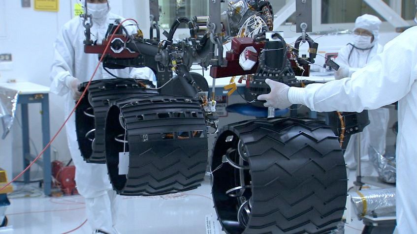 In the middle of this image, three wheels are shown raised by a lift, with engineers on both sides of the wheels in the cleanroom, where the Curiosity rover is being assembled. The wheels are made out of aluminum but have a black coating, which prevents the wheels from slipping.  The wheels are attached to dark poles, which bend like 'joints.'  Several black and red cable wires are visible coming out of the poles.  An engineer stands in the background holding a clipboard and looking closely at the rover.  A desk and various tools are visible behind the wheels on the right.