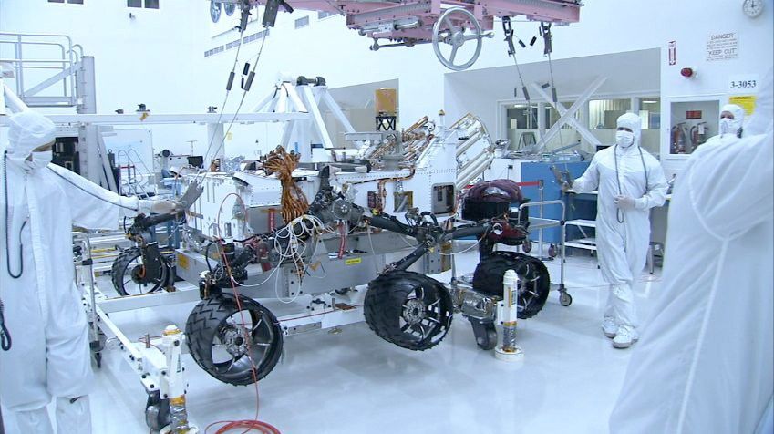 In this picture, the Curiosity rover is sitting on top of six shiny wheels.  The picture was taken from the side of the rover and only four wheels are visible.  The wheels have a black coating which makes them slip resistant.  On the right of the rover is an engineer who is wearing a white 'bunny suit' to prevent him from getting any unwanted Earth microbes onto the rover.  The engineer is holding one of the lift cables used to put the rover onto the wheels.  A pink heavy-lift crane is visible over the rover.  The crane is used to lift the rover body within the room.  Behind the rover, lots of lab equipment and other parts are scattered around the cleanroom.