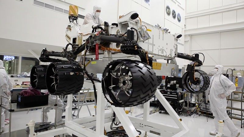 This image was taken in the clean room where the Curiosity rover is being assembled.  It shows the rover, which is about the size of an SUV, hoisted on a white lift, with its black wheels suspended in the air.   One engineer is on top of the hoist and is leaning over the rover body, while another is looking up on the ground floor to the right of the rover.  Both engineers are wearing white "bunny suits" to keep them from contaminating any equipment.