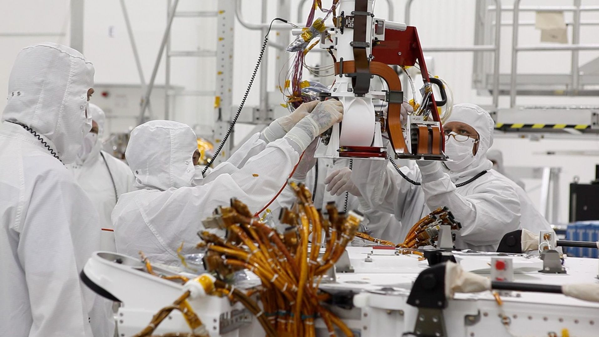In this image, engineers are dressed head to toe in "bunny suits" (white hoods, lab-style coats and gloves).  Only their eyes and foreheads can be seen.  They are huddled around the base of the rover's "neck" (its Mast).  They watch intently as they carefully lower the Mast to attach it to the rover's flat "back."  A cluster of yellow and red wires on the rover's body pokes up in the foreground of the image.