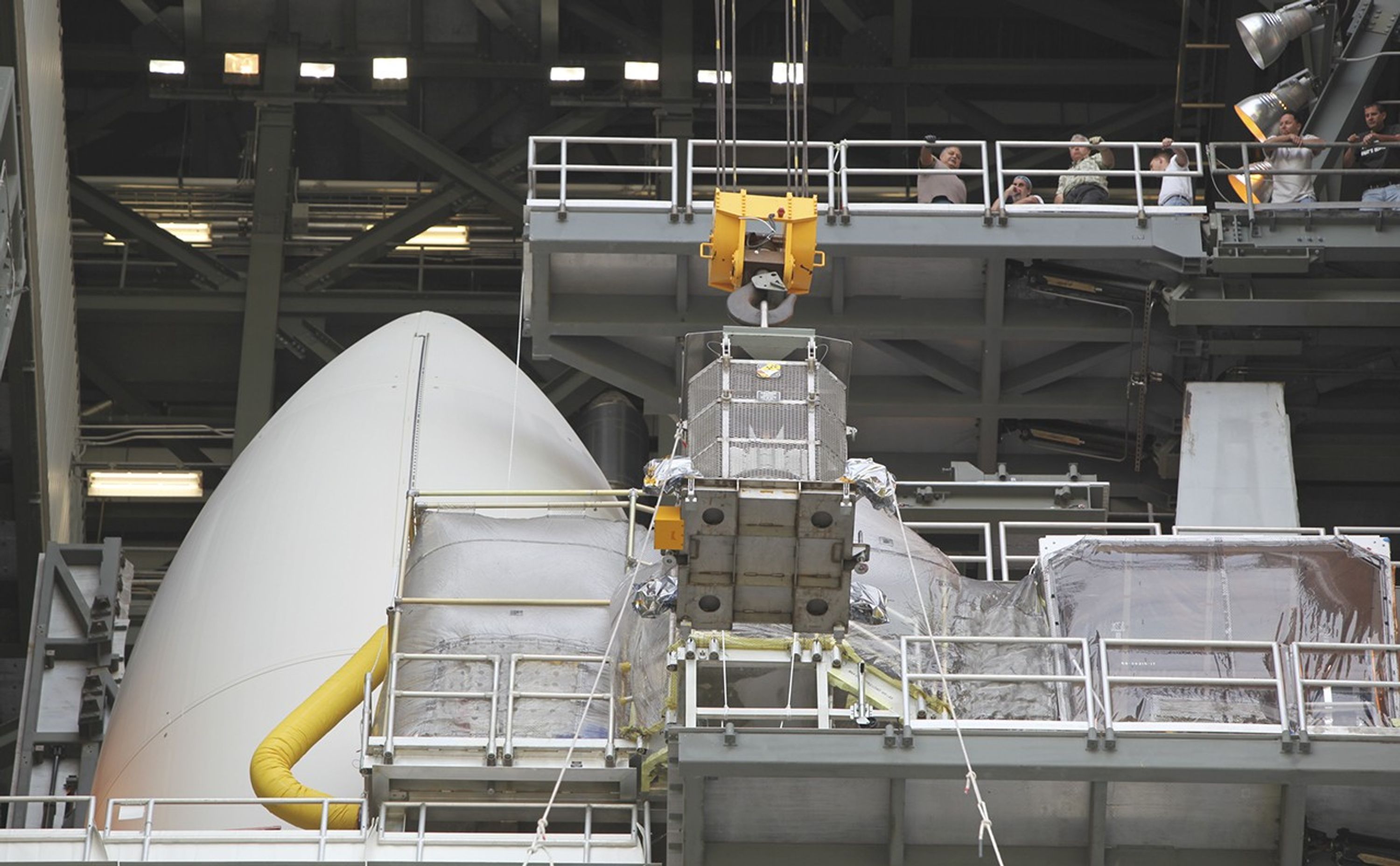 Employees gathered one level above monitor the progress of the protective mesh container known as the "gorilla cage," holding the multi-mission radioisotope thermoelectric generator (MMRTG) for NASA's Mars Science Laboratory (MSL) mission, as it is lifted near the top of the Atlas V rocket in the Vertical Integration Facility at Space Launch Complex 41.