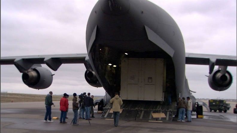 In this image, more than a dozen people stand, in winter coats, behind a huge, gray military C-17 cargo plane.  From this vantage point, the plane resembles a huge shark with its mouth wide open.  Inside the 'mouth' is one of the large boxes that contain the Mars Reconnaissance Orbiter.  The wings of the plane jut out from both sides.  The sky is cloud-covered.