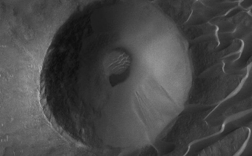 The dramatic MOC narrow angle camera image presented here was acquired in June 2006. It shows a crater that has been encroached by a field of dark, windblown sand dunes in the Syrtis Major volcanic region of Mars. The area downwind of the crater (to the left/lower left) is free of dunes because the raised rim of the crater prevented winds from causing sand to be deposited in the crater's lee.