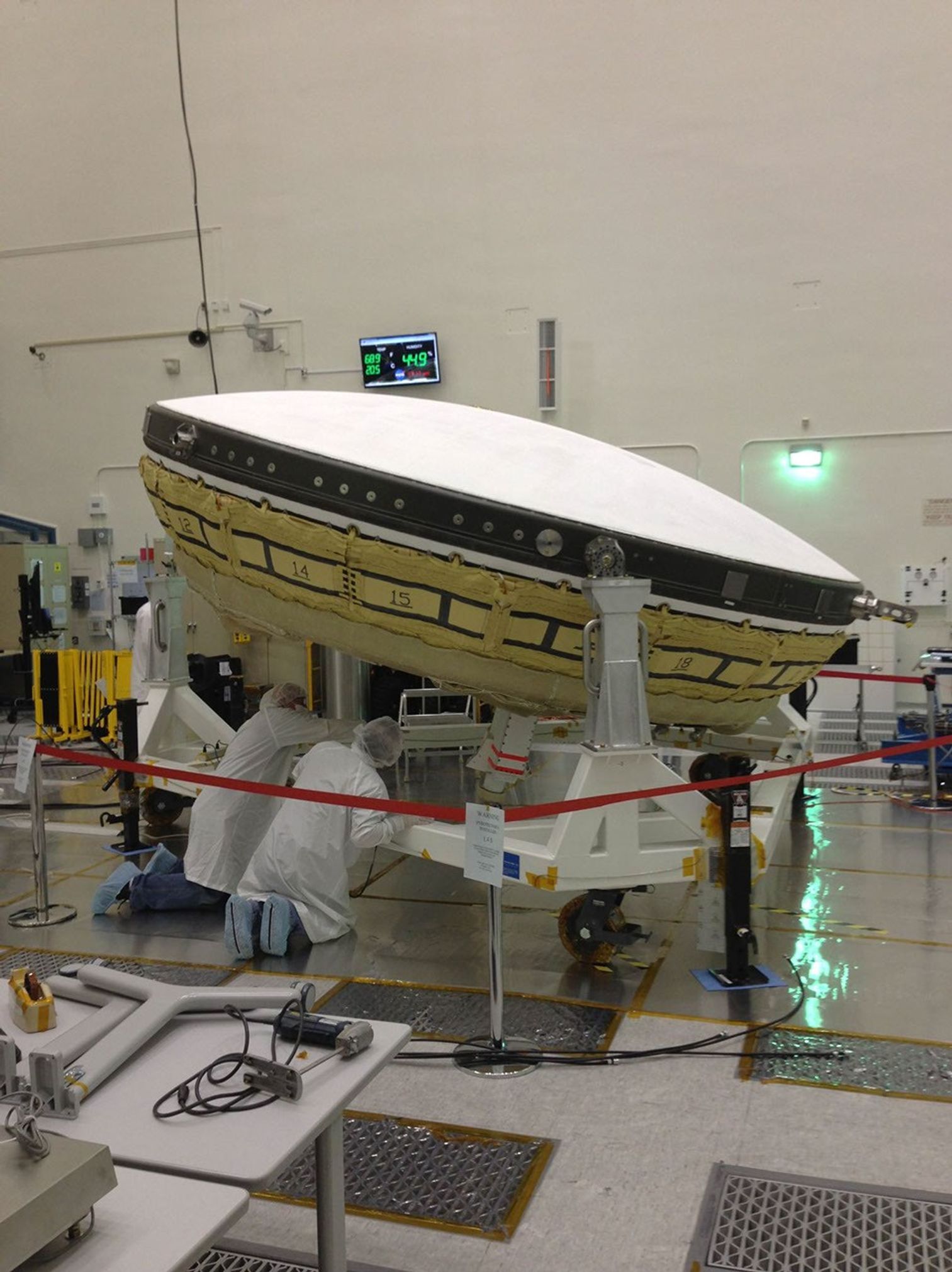 NASA workers at JPL wearing clean room, white "bunny suits," prepare a large saucer-shaped test vehicle that will be used to land larger payloads on Mars.