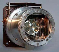 This photo shows a round, metal cylinder, small enough to fit in a human palm, open at  the end to show a flat bottomed-interior with a smaller, circular metal plate at the bottom. Around the edges of the circular metal plate are six circular openings arranged in a daisy-like formation, like the chamber openings at the end of a six-cylinder pistol.