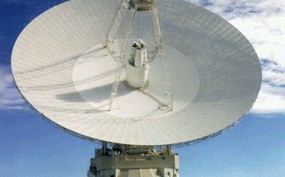 The image shows a very tall, wide, dish-shaped device that is about three times as wide and as tall as the two-story dome that supports it. Four metal ladderlike extensions form a pyramidal structure topped by a box-like frame used for transmission and reception. The entire array is pointed skyward. In the background is a partly cloudy, blue sky above sagebrush-covered desert hills.