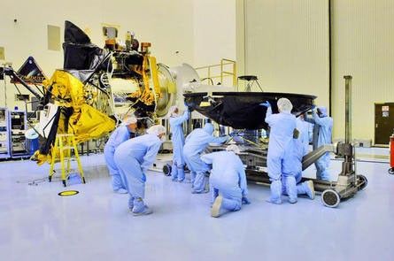 Technicians and engineers dressed from head to toe in blue coveralls (called 'bunny suits') prepare the Mars Reconnaissance Orbiter's large, black high-gain antenna (that resembles a satellite dish) to be mated with the body of the spacecraft.  The bus of the spacecraft with nearly all of its instruments in place, sits just to the left of the people pictured.  Parts of the orbiter are covered in gold, shiny thermal blanketing.  Facing directly upward is the spacecraft's huge Hi-RISE camera covered in black protective blanketing.