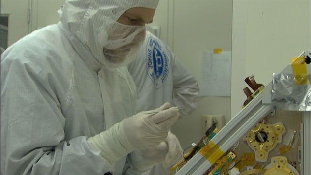 In this image, a Caucasian man wears a white, protective suit in a cleanroom. His face is mostly covered and his hands are gloved. He is working on the CRISM instrument for the Mars Reconnaissance Orbiter mission. The instrument sits on a slanted aluminum-like board covered with yellow Kapton tape - a low-static tape safe for using in cleanrooms and near or on instrumentation.