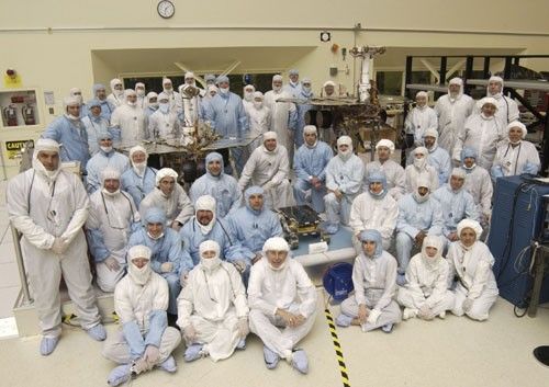 A team of about 50 men and women in white and blue cleanroom smocks and bonnets stand and sit around their creations: Spirit and Opportunity. The twin rovers of the Mars Exploration Rover mission are about the size of golf carts. The significantly smaller, microwave-sized Sojourner rover from the 1997 Pathfinder mission sits in the middle.